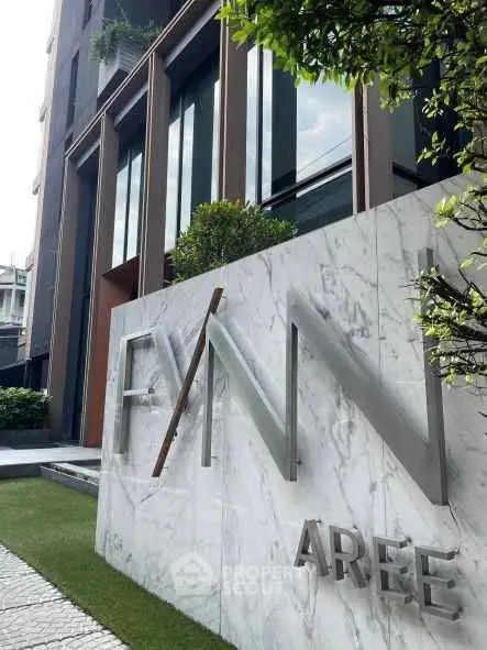 Modern building entrance with sleek design and stylish signage, surrounded by lush greenery.