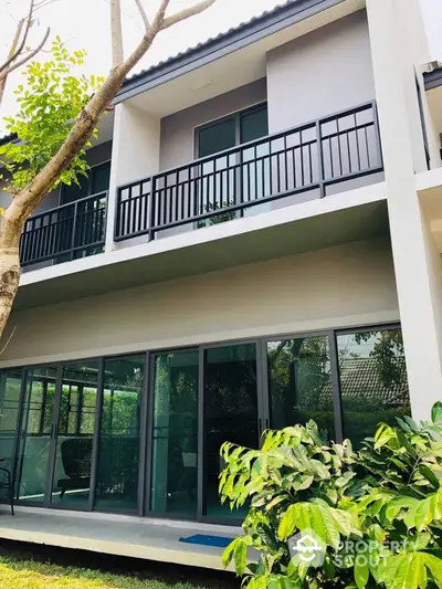 Modern two-story house with balcony and large glass doors in lush garden setting.