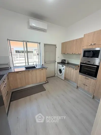 Modern kitchen with wooden cabinets, built-in oven, and washing machine in a bright, airy space.