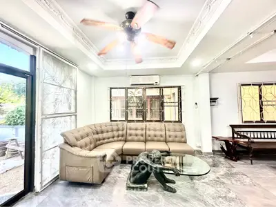 Spacious living room with elegant sofa and unique glass coffee table, featuring a ceiling fan and large windows.