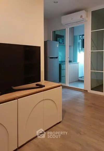 Modern living room with sleek TV stand and view into a compact kitchen with fridge and air conditioning.