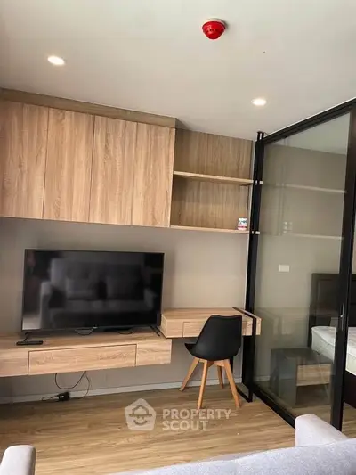 Modern living room with sleek wooden furniture and glass partition, featuring a mounted TV and cozy workspace.
