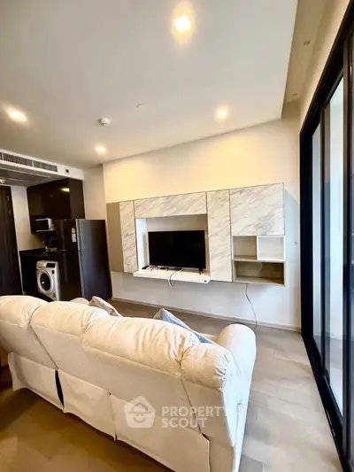 Modern living room with sleek design, featuring a cozy sofa, built-in TV unit, and compact kitchen area with appliances.