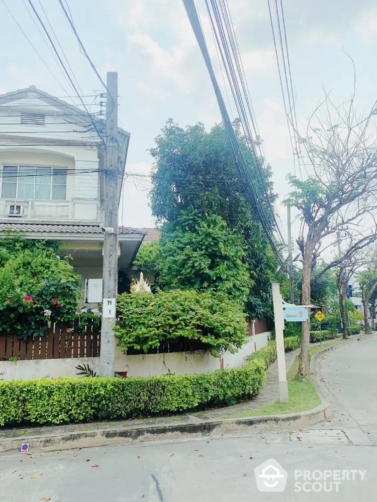 Charming two-story residence nestled in a lush, green neighborhood, boasting a well-manicured hedge and vibrant flora, complemented by a serene, tree-lined street.