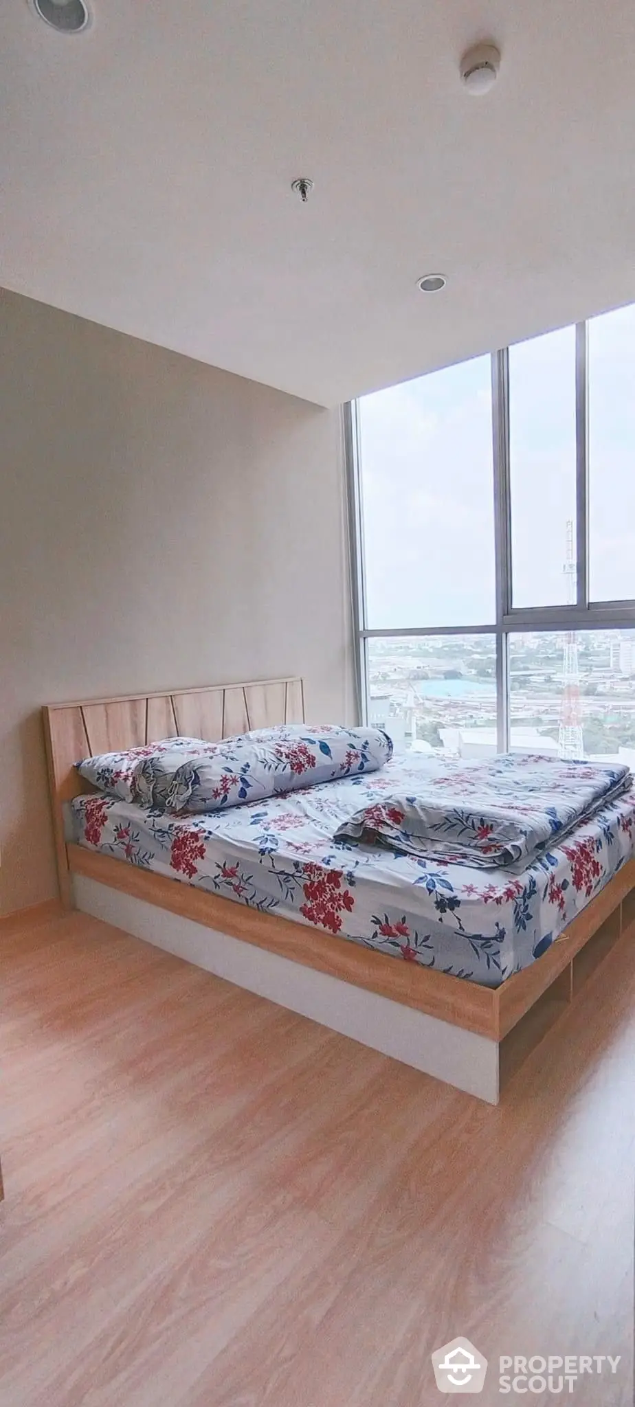 Spacious bedroom with large windows offering ample natural light and a stunning city view, featuring a modern wooden bed with a stylish floral bedding set.