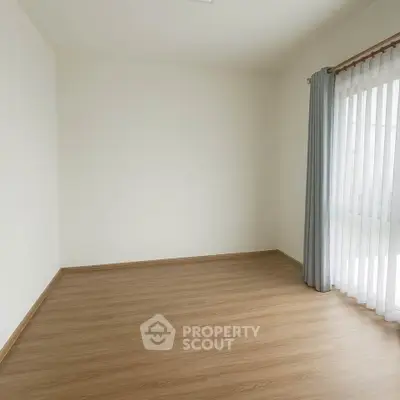 Spacious empty room with wooden flooring and large window with curtains.