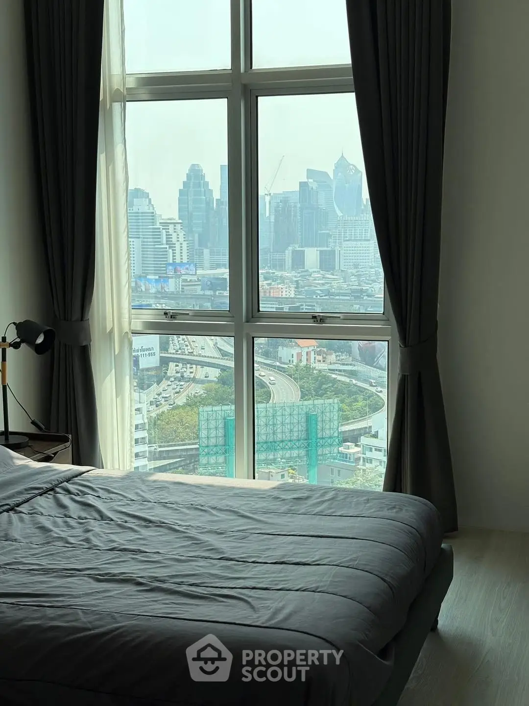 Modern bedroom with stunning city view through large windows, featuring cozy bedding and elegant curtains.