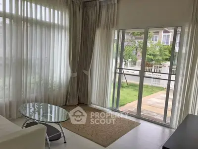 Bright living room with large windows and garden view, featuring modern glass coffee table and elegant curtains.