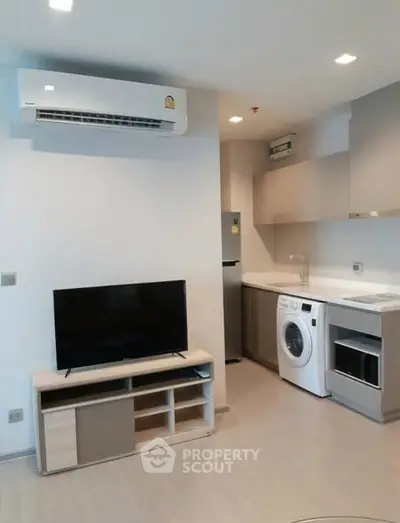 Modern apartment interior with air conditioning, TV, and compact kitchen featuring washing machine and fridge.