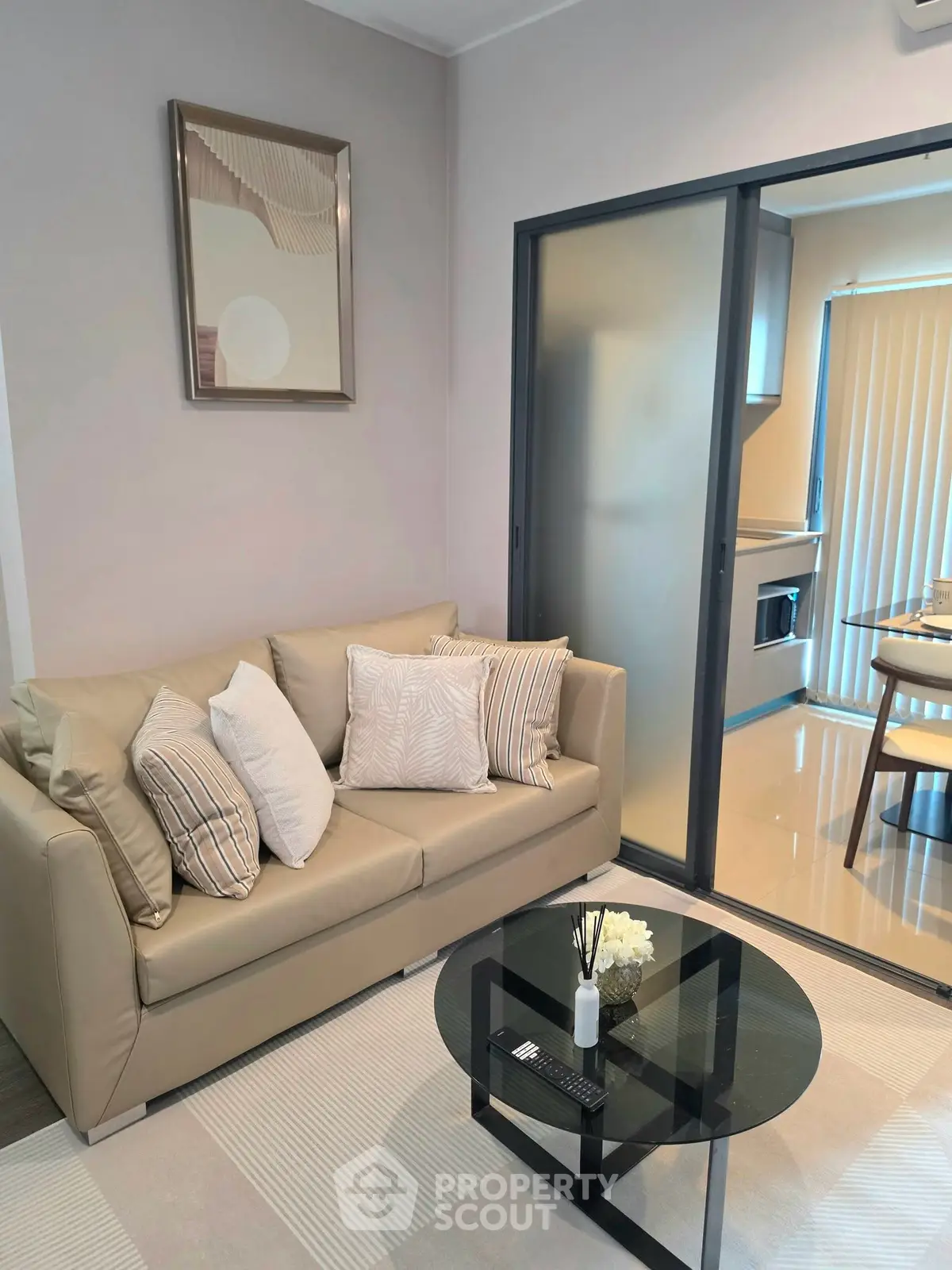 Modern living room with beige sofa and glass coffee table, adjacent to a sleek kitchen area.