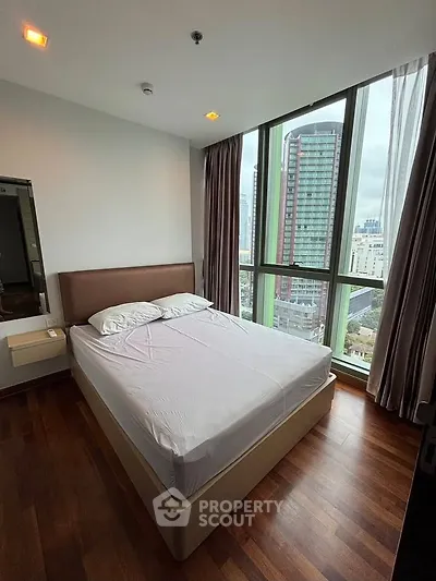 Modern bedroom with large windows offering city views, featuring a comfortable bed and wooden flooring.