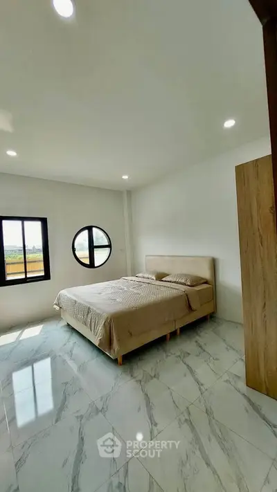 Modern bedroom with large windows and circular window, featuring a cozy bed and sleek marble flooring.