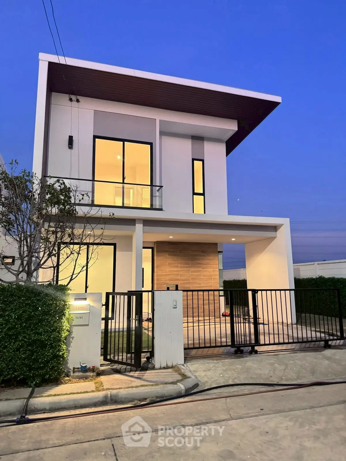 Modern two-story house with sleek design and large windows, featuring a gated entrance and manicured hedges.