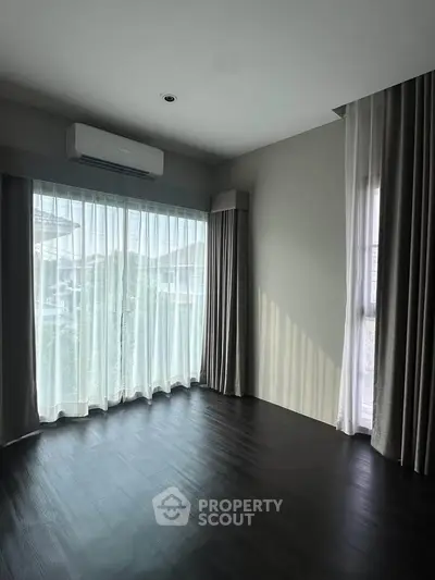 Spacious living room with large windows and elegant curtains, featuring dark wood flooring and modern air conditioning.