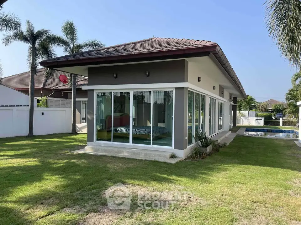 Modern single-story house with large windows and lush garden, featuring a private pool and palm trees.
