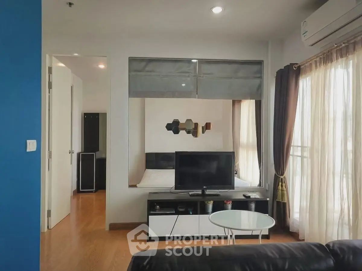 Modern living room with TV, glass table, and view into bedroom through interior window.