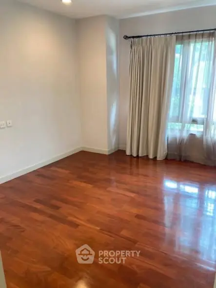 Spacious empty room with polished wooden floors and large window with curtains.