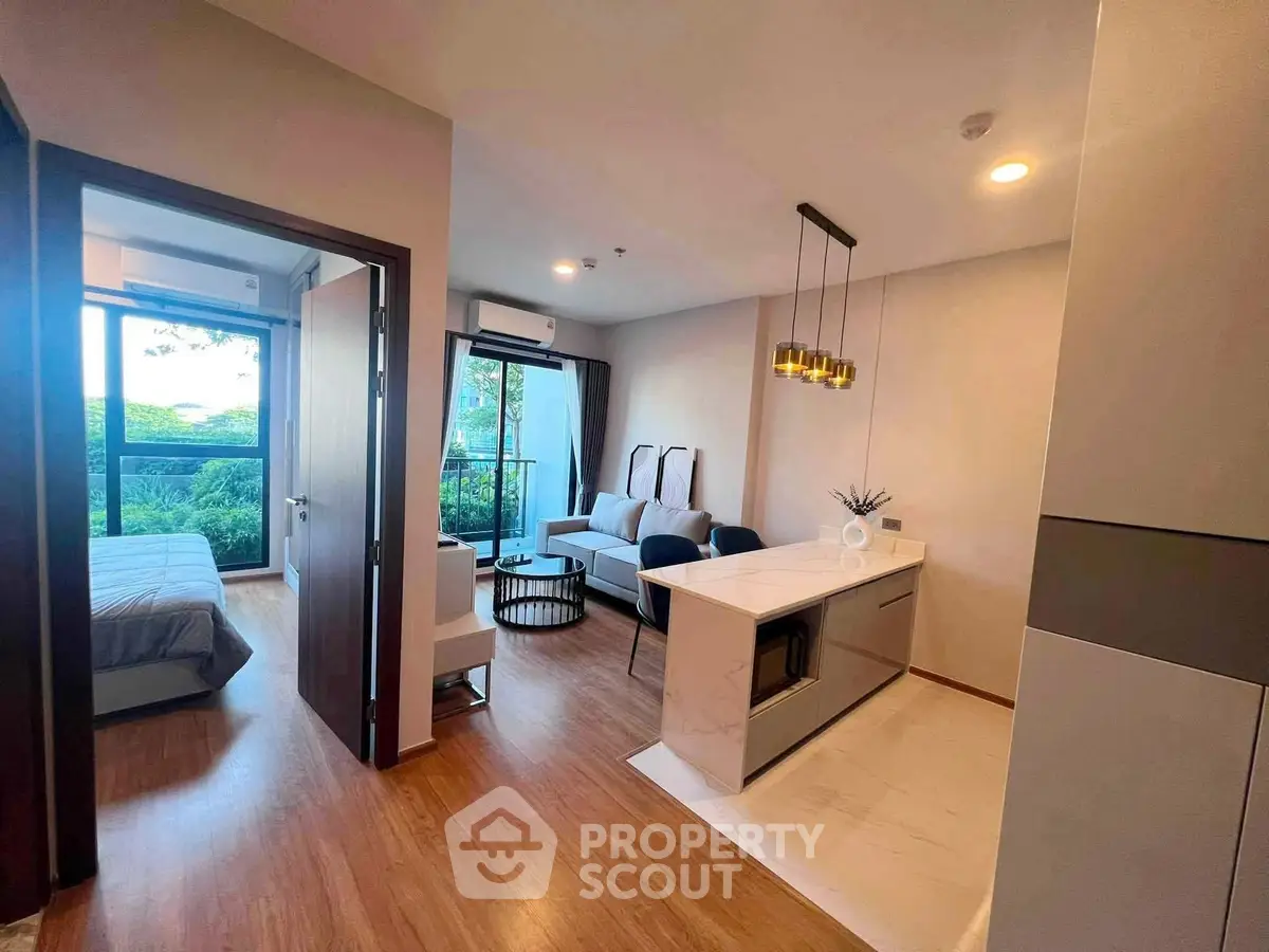 Modern open-plan living room with kitchen and bedroom, featuring elegant lighting and large windows with a garden view.