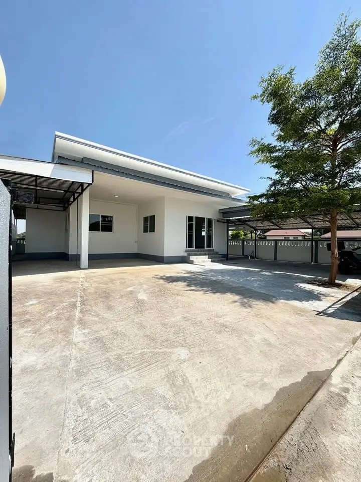 Modern single-story house with spacious driveway and carport, surrounded by greenery under a clear blue sky.