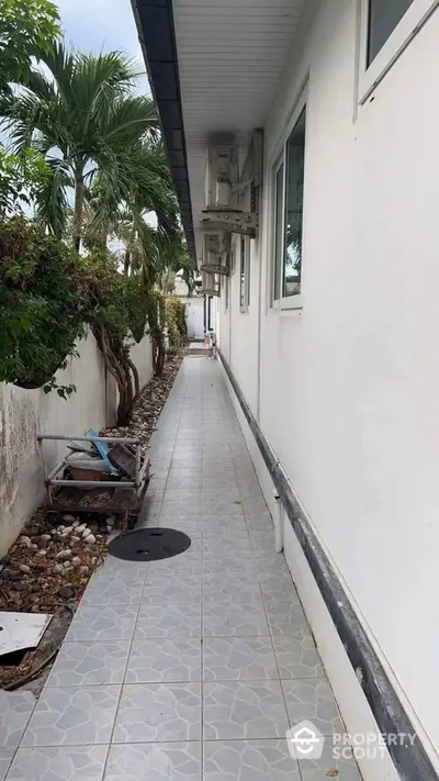 Narrow exterior walkway beside a white building with tiled floor and lush greenery.