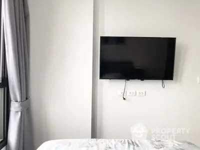 Modern minimalist bedroom with a wall-mounted flat-screen TV, crisp white walls, and natural light filtering through sheer curtains.