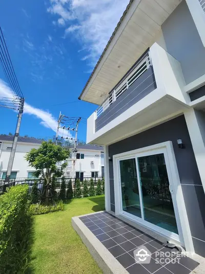 Modern two-story house with balcony and lush green garden under clear blue sky.