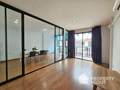 Spacious and modern living room with large glass windows offering ample natural light, sleek wooden flooring, and a seamless transition to a cozy dining area.