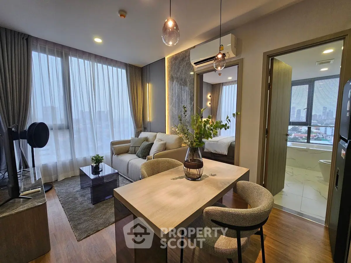 Modern living room with dining area and view into bedroom, featuring stylish decor and large windows.