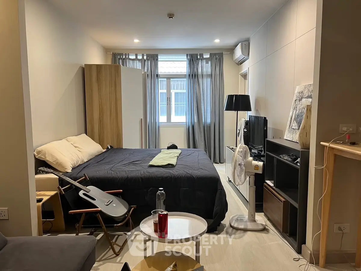 Modern studio apartment with cozy bed, stylish decor, and natural light from large window.