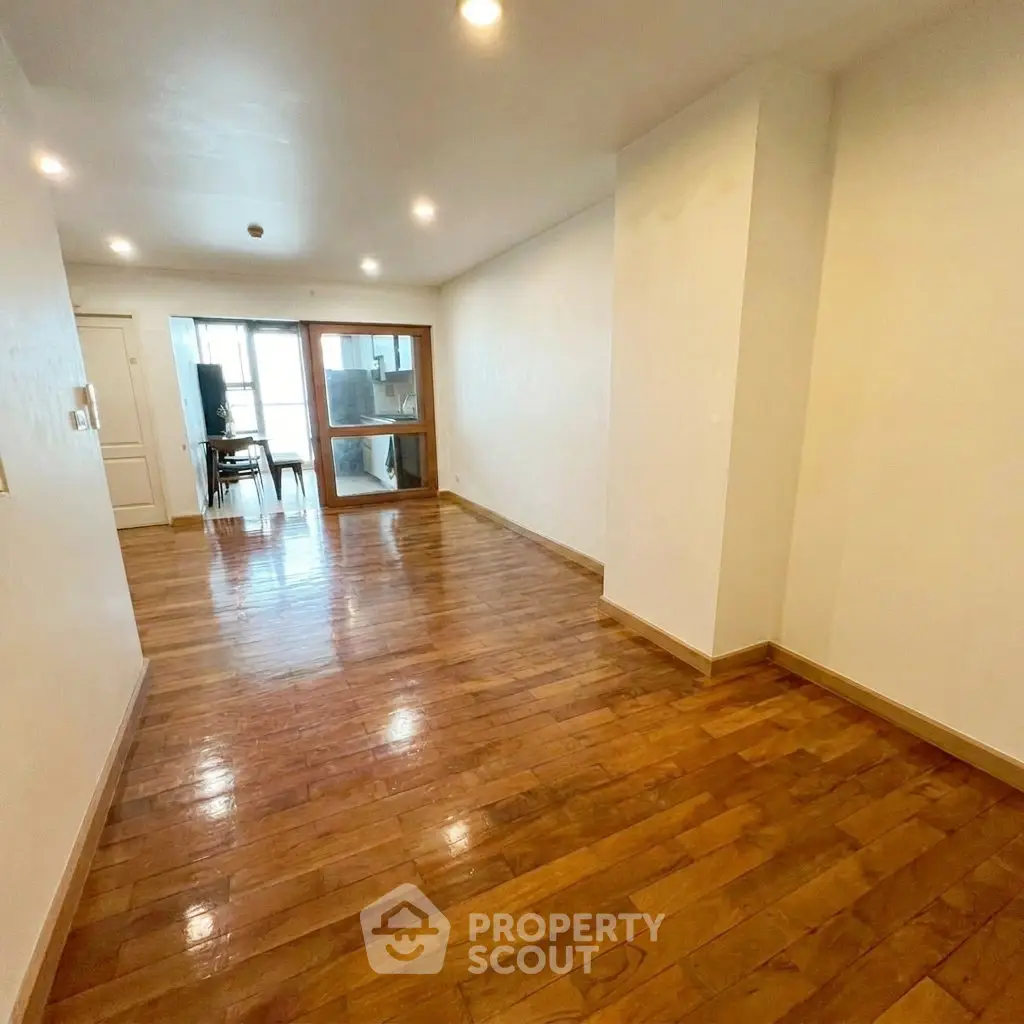 Spacious hardwood-floored living room with bright lighting and glass door leading to another area.