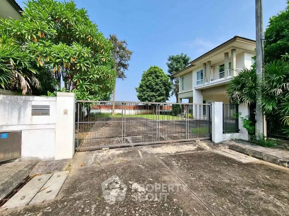 Spacious gated entrance to a modern two-story house with lush greenery and ample driveway space.