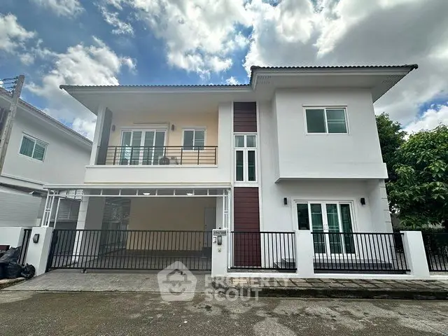 Modern two-story white house with balcony and gated driveway under a partly cloudy sky.