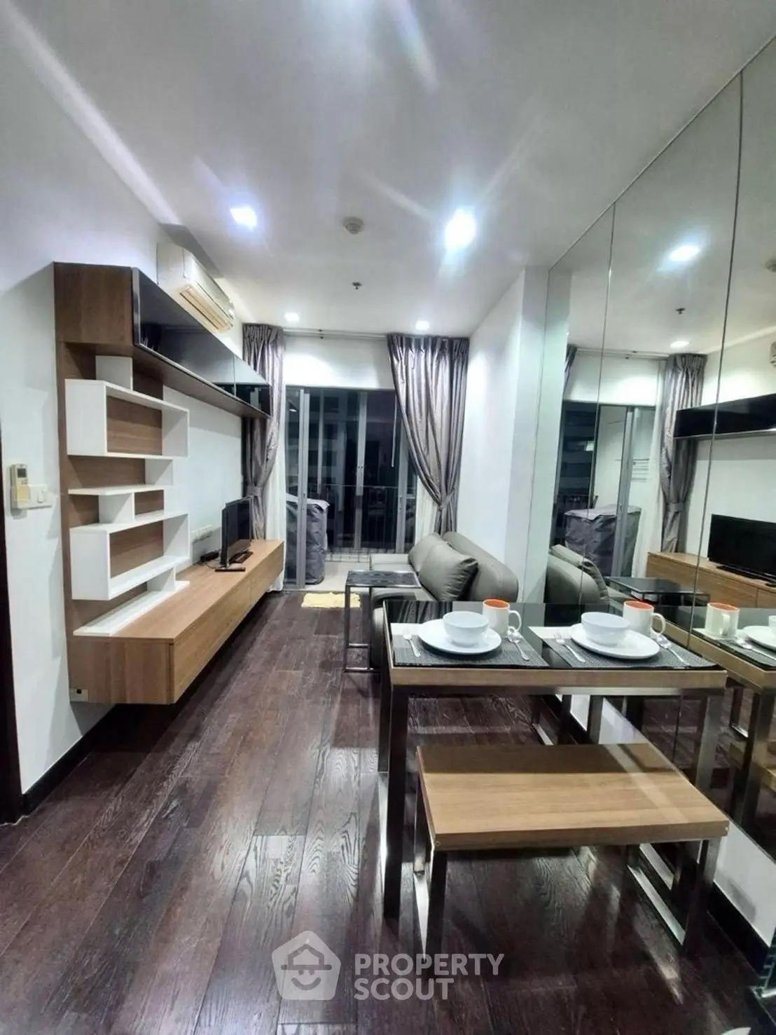 Modern living room with sleek furniture and dining area, featuring dark wood flooring and mirrored wall.