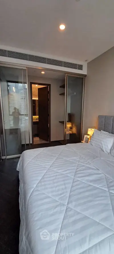 Modern bedroom with cozy bedding and city view, featuring sleek glass doors and ambient lighting.