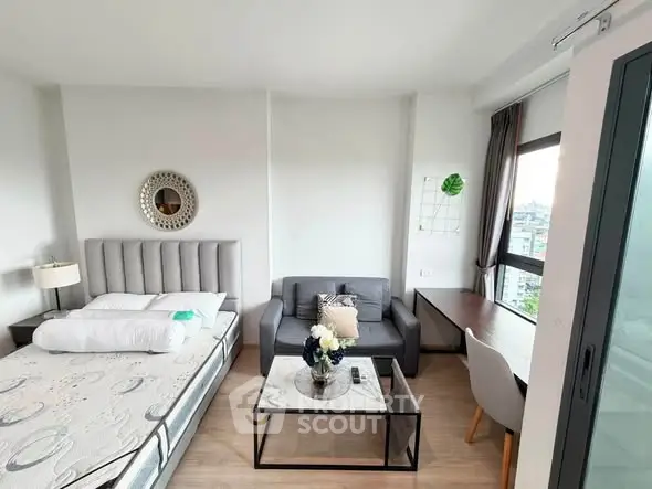 Modern studio apartment with bed, sofa, and desk, featuring large window with city view.