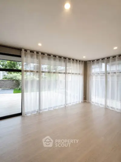 Spacious empty living room with large windows and sheer curtains, offering abundant natural light and garden view.