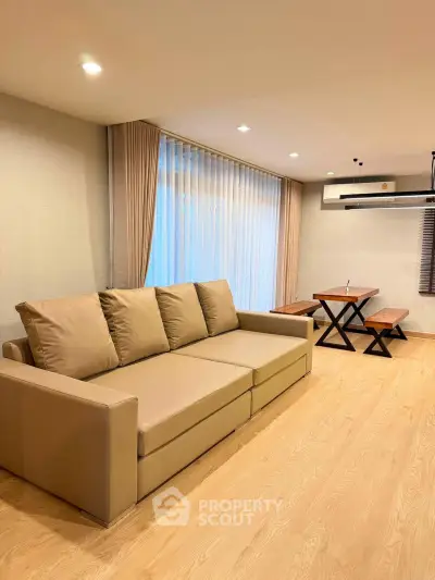 Modern living room with beige sofa and dining area, featuring wooden flooring and large windows.