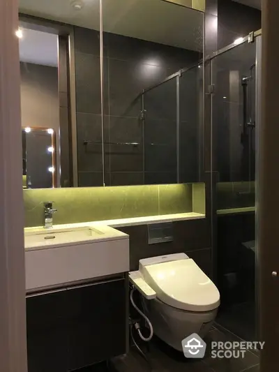 Modern bathroom with sleek dark tiles, underlit green vanity, large mirror, and glass-enclosed shower, exuding sophistication and style.