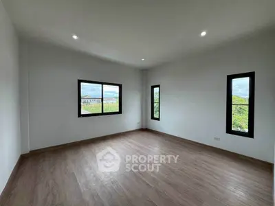 Spacious empty room with large windows and wooden flooring, offering a serene view of greenery.