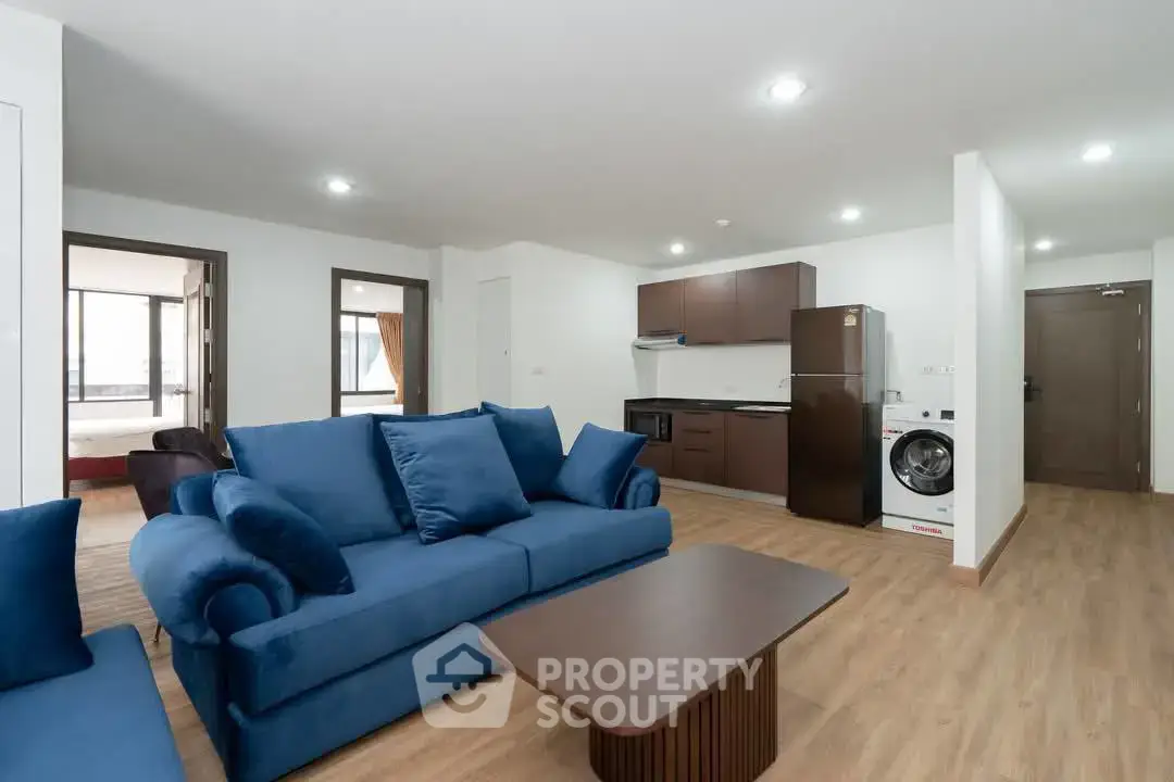 Spacious modern living room with blue sofa and open kitchen layout, featuring a washing machine and fridge.