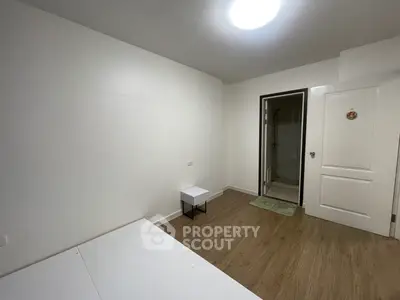 Minimalist bedroom with wooden flooring and white walls, featuring a small table and a door leading to an ensuite bathroom.