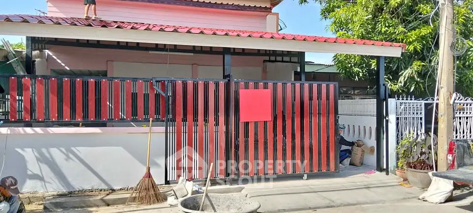 Charming suburban home with red and black gate, lush greenery, and spacious driveway.