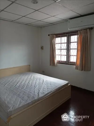 Cozy bedroom with polished wooden floors and natural light filtering through traditional window curtains, complemented by a large comfortable bed and an air conditioning unit for optimal climate control.