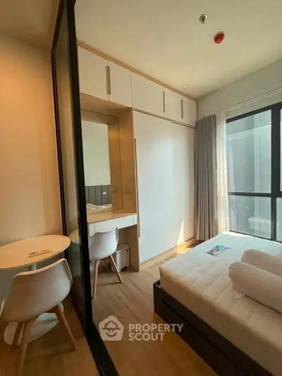 Modern bedroom with natural light, featuring a cozy bed, built-in wardrobe, and a small dining area.