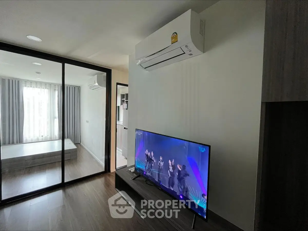 Modern living room with sleek design, featuring a wall-mounted air conditioner and a flat-screen TV, adjacent to a glass-walled bedroom.