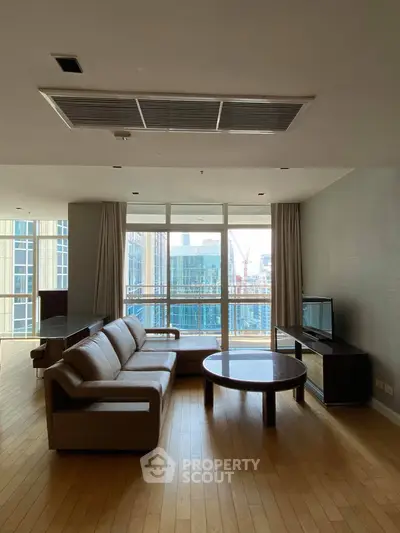 Spacious living room with modern furniture and city view, featuring large windows and wooden flooring.