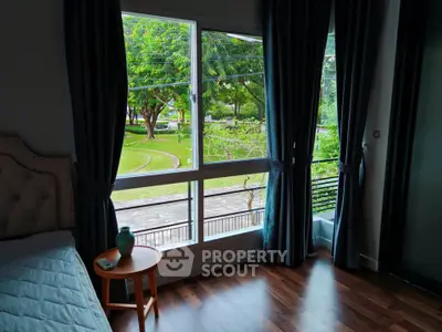 Cozy bedroom with large window offering a serene garden view, featuring elegant curtains and wooden flooring.
