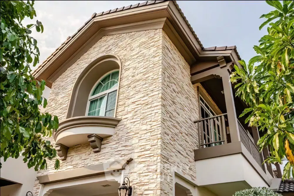 Charming two-story house with stone facade and elegant balcony surrounded by lush greenery.