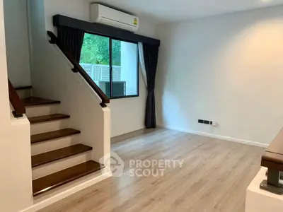 Modern living room with wooden flooring and staircase, featuring large window and air conditioning.