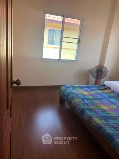 Cozy bedroom with wooden flooring and colorful bedding, featuring a window with a view of the neighboring building.